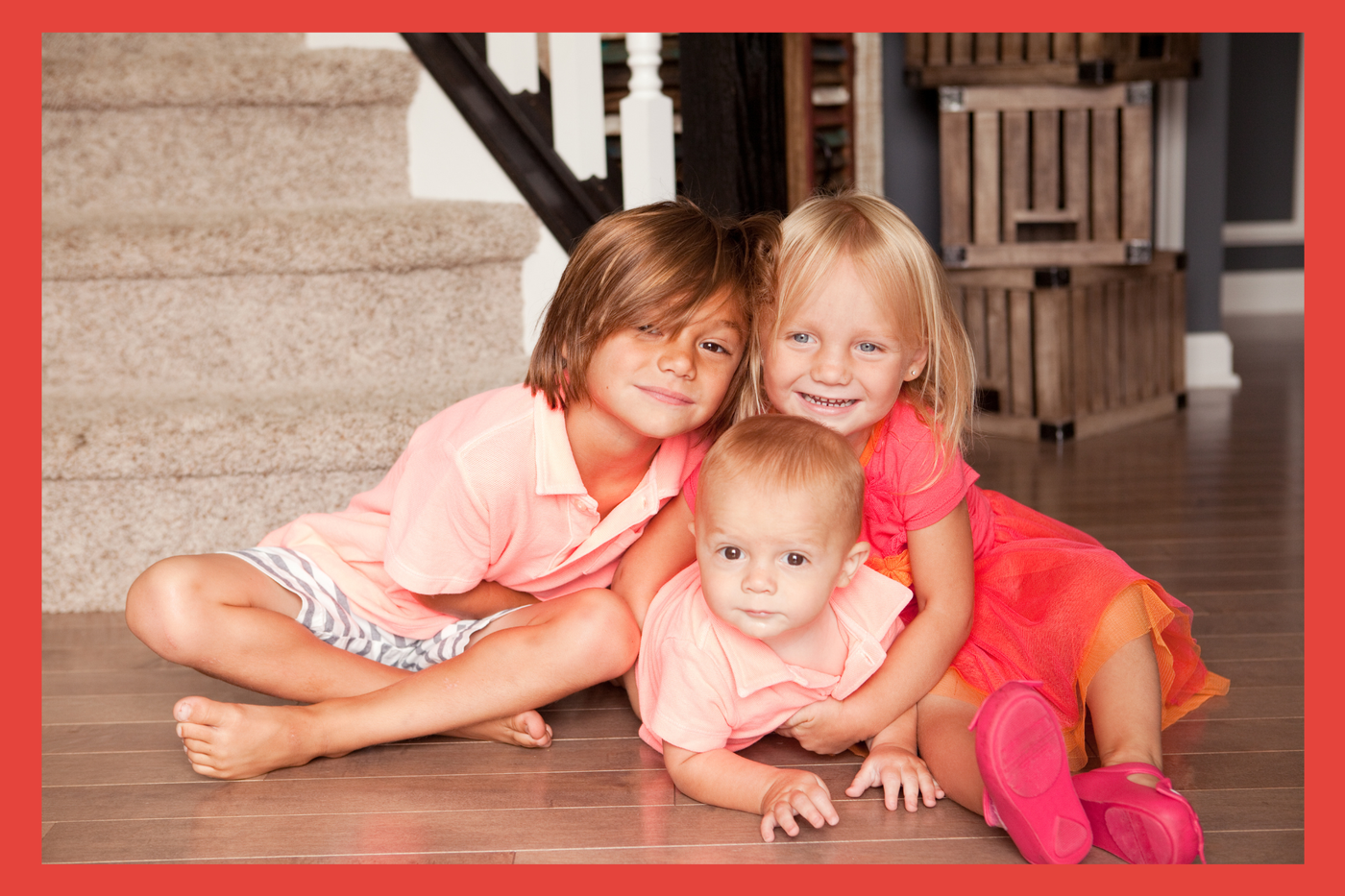 Toddler boy with longer hair in shorts and polo. Toddler girl in a pink tye die dress with blonde hair. Baby boy in a pink polo. All hugging and posed in front of the stairs. 